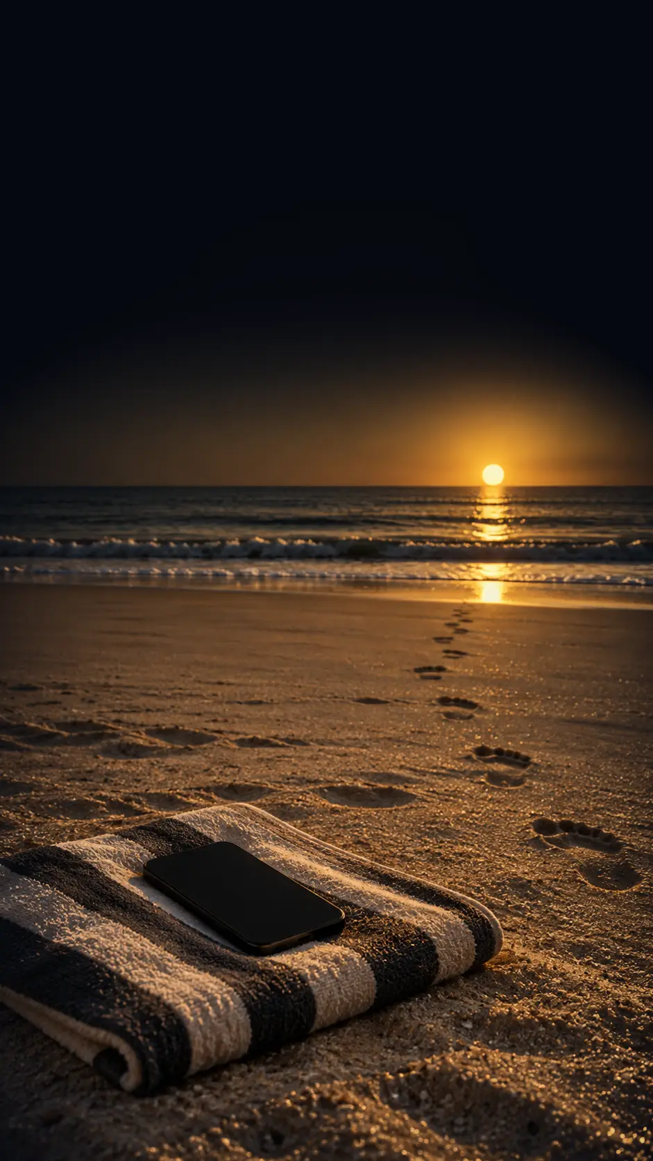 A towel on the sand at sunset, a phone lying on it, footprints to the water